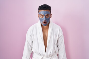 Canvas Print - Young hispanic man wearing beauty face mask and bath robe afraid and shocked with surprise and amazed expression, fear and excited face.