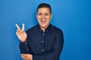 Canvas Print - Hispanic young man standing over blue background smiling with happy face winking at the camera doing victory sign. number two.