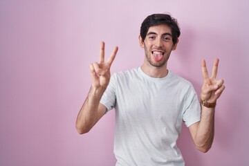 Wall Mural - Young hispanic man standing over pink background smiling with tongue out showing fingers of both hands doing victory sign. number two.