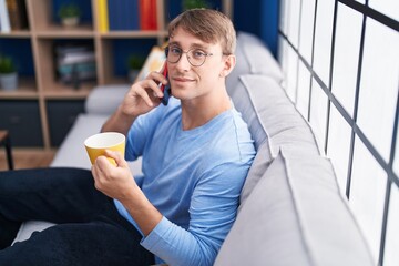 Wall Mural - Young caucasian man talking on smartphone drinking coffee at home