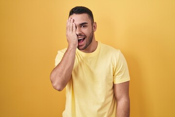 Canvas Print - Young hispanic man standing over yellow background covering one eye with hand, confident smile on face and surprise emotion.