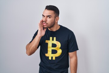 Poster - Young hispanic man wearing bitcoin t shirt hand on mouth telling secret rumor, whispering malicious talk conversation