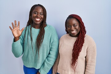 Sticker - Two african woman standing over blue background showing and pointing up with fingers number five while smiling confident and happy.