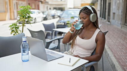 Canvas Print - African american woman using laptop and headphones drinking glass of water at coffee shop terrace