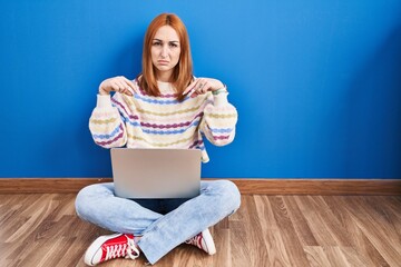 Sticker - Young woman using laptop at home sitting on the floor pointing down looking sad and upset, indicating direction with fingers, unhappy and depressed.