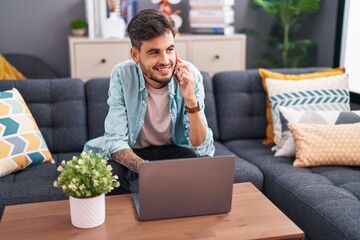 Wall Mural - Young hispanic man talking on smartphone using laptop at home