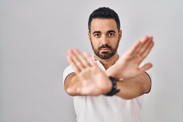 Poster - Young hispanic man with beard wearing casual clothes over white background rejection expression crossing arms and palms doing negative sign, angry face