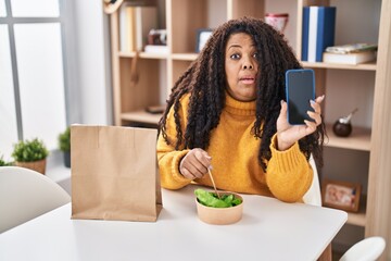 Sticker - Plus size hispanic woman eating take away salad holding smartphone clueless and confused expression. doubt concept.