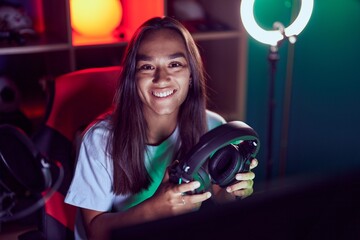 Wall Mural - Young beautiful hispanic woman streamer smiling confident holding headphones at gaming room