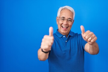 Sticker - Middle age man with grey hair standing over blue background approving doing positive gesture with hand, thumbs up smiling and happy for success. winner gesture.