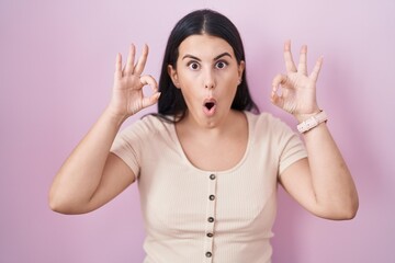 Canvas Print - Young hispanic woman standing over pink background looking surprised and shocked doing ok approval symbol with fingers. crazy expression