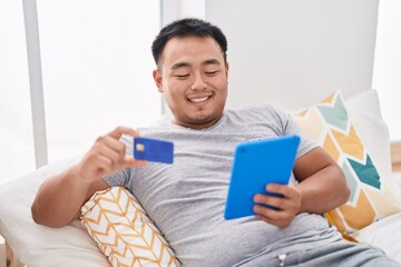 Wall Mural - Young chinese man using touchpad and credit card sitting on bed at bedroom