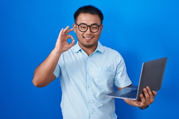 Canvas Print - Chinese young man using computer laptop smiling positive doing ok sign with hand and fingers. successful expression.