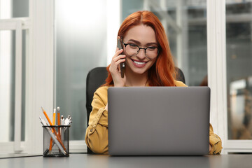 Poster - Happy woman talking on smartphone while working with laptop at desk in office