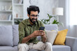 © Liubomir - Young smiling man happy sitting at home on the sofa in the living room, Indian man uses an application on the phone, dials a message, calls, and browses online sites on the Internet.