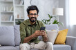 © Liubomir - Portrait of happy man alone at home, hispanic man smiling and looking at camera, holding phone while sitting on sofa in living room of apartment house.
