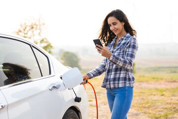 World Environment Day. save world save life. Hispanic female driver electric car holding electrical plug connector to charge the batteries hybrid car at home or charging station. Using smartphone