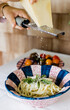 woman's hands grating a block of fresh pecorino romano cheese onto a purple and blue ceramic bowl of fresh pasta with heirloom tomatoes in background