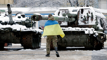 
A man with a Ukrainian flag in front of a battle tank. Manifestation of protest against the invasion of Russia, the war in Ukraine. A man wrapped in a flag stands next to a military vehicle, an anti-