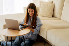 Happy Young Woman Reading Book While Sitting With Laptop Near Sofa