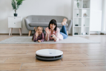 Focus on robot vacuum carrying out scheduled cleaning task while female owners waving goodbye from behind. Smart device saving energy from tedious home cleaning work by using advanced technologies.