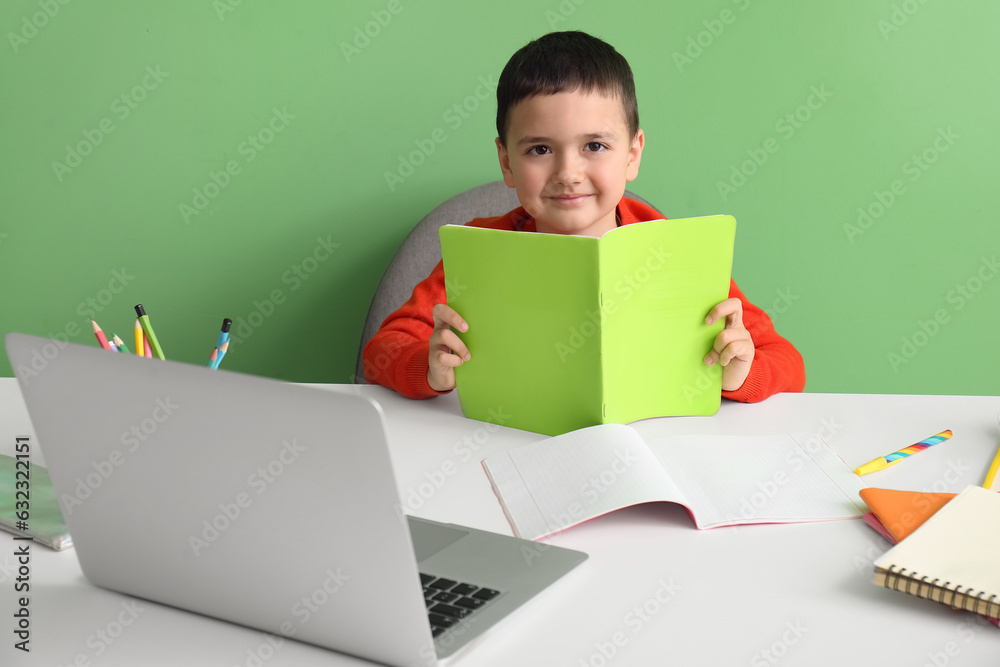 Cute little boy with copybook doing homework at table near green wall