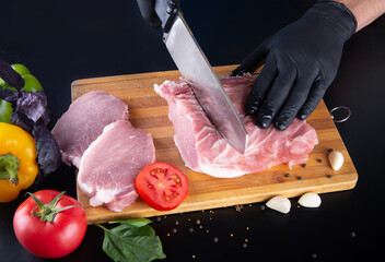 Chief cooking juicy beef steak on dark black background. Man hands in black gloves cutting meat on wooden board, fresh vegetables, garlic, basil leaves, pepper seeds. Restaurant menu, recipe concept