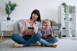 © sofiko14 - Close up view of happy mom and tween girl holding gaming consoles while laughing on carpet in modern apartment. Playful adult and child embracing challenges of virtual world at leisure time.