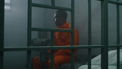 Wall Mural - African American prisoner in orange uniform sits on the bed behind bars, reads Bible in prison cell. Criminal serves imprisonment term for crime in jail. Detention center or correctional facility.