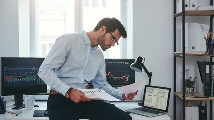 Fototapete - Busy working day. Young successful trader or businessman in formal wear and eyeglasses sitting at modern office in front of computer screens with charts and reading market report