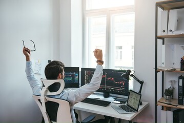 Fototapete - Winner Back view of stock trader with raised hands holding eyeglasses and feeling happy while looking at multiple computer screens with data and charts in his modern office.