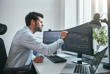 Fototapete - Analyzing data. Thoughtful young trader is pointing at graphs on computer screen while working in his modern office.