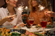 © Seventyfour - Close-up of happy young friends holding sparklers over served table with homemade food and snacks and two glasses of red wine at party