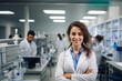 © uv_group - Beautiful young woman scientist wearing white coat and glasses in modern Medical Science Laboratory with Team of Specialists on background.