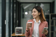 © NINENII - Young woman standing with coffee cup and smartphone, thinking and looking out window during working day