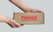 One rectangular cardboard box is held in the hands on a plain light background. A box with an adhesive warning label for packaging - Fragile, Handle with care, Thank you. No people. Corner composition