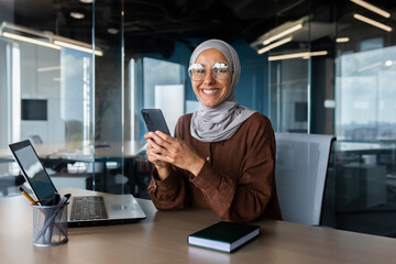 Wall Mural - Smiling young Muslim woman in hijab working in office on laptop, using mobile phone, smiling at camera.