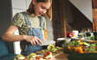 © Ievgen Skrypko - Girl child cuts avicado with knife at kitchen. Pretty female kid with fruits and vegetables preparing salad