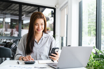 Canvas Print - pretty happy young asian entrepreneur woman holding mobile phone working with laptop sitting in the office or co working space cafe.