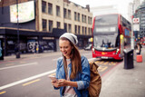 Fototapeta Londyn - Young woman using a smart phone while waiting for her bus at a bus stop in London