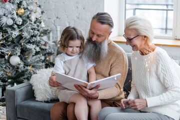 Senior grandparents, attractive grandmother and handsome grandfather reading a fairy tale book to cute little granddaughter near decorated Christmas tree. Two generations, family values, happy people