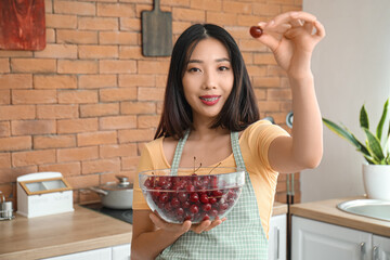 Wall Mural - Happy beautiful young Asian woman in apron with bowl of ripe cherries in kitchen