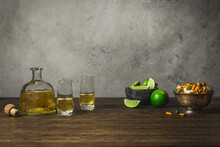 Tequila Bottle And Shot Glasses With Limes And Small Bowl Of Spicy Rice Crackers.