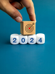 Poster - The calendar year 2024 with goal and success concept. Target icon on wooden cube put by hand on white blocks with 2024 number, isolated on blue background. Happy new year, big business goals banner.