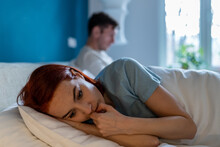 Upset Wife Lies With Her Back To Husband. Frustration, Unhappiness, Pain, Resentment And Thoughtfulness On Face Of Sad Depressed Woman Lying In Bed Thinking. Man With Disinterested Look Ignores Woman.