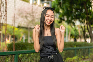 A pretty lady standing in the park wearing a black strapless tank top and sunglasses, smiling widely with closed eyes and holding up two closed fists. A building trees and bushes in the background.