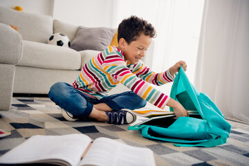 Wall Mural - Photo of cute little boy sitting floor in living room flat prepare school bag for tomorrow day put book notebook