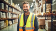 © BlazingDesigns - Smiling portrait of handsome a warehouse worker in protective vest. Background stock of parcels with products ready for shipment.
