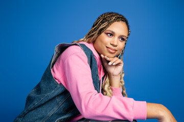 Wall Mural - charming and dreamy dark skinned woman in denim vest and hoodie looking away on blue background