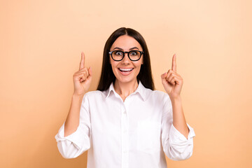 Wall Mural - Photo of sweet excited woman dressed white shirt eyewear pointing two fingers up empty space isolated beige color background